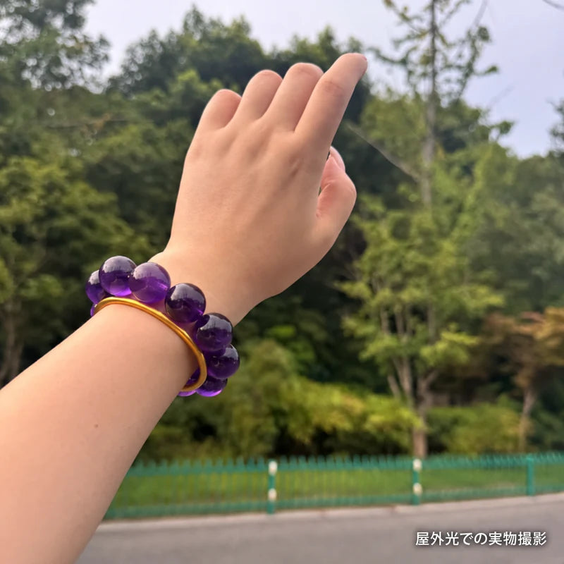 緑の木々を背景にゴールドカラーのバングルと合わせたウルグアイアメジストブレスレット。屋外光で撮影した実物写真